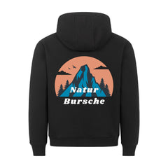 Der MarketPrint Premium Hoodie "Natur Bursche" in schwarz hat eine bequeme Passform und eine blaue Berg-, Tannenbaum-, Vogel- und Wolkengrafik auf einem orangefarbenen Kreis mit "Natur Bursche" in weiß. Hergestellt in Türkiye.