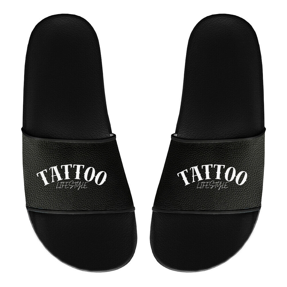 Ein Paar schwarze MarketPrint Badelatschen "Tattoo" aus PVC mit dem Schriftzug "TATTOO LIFESTYLE" in weiß auf den Trägern, die von oben auf einem schlichten weißen Hintergrund zu sehen sind.