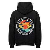 Der MarketPrint Oversized Hoodie "Urlaub" ist schwarz mit einem auffälligen, kreisförmigen Sonnenuntergang-, Meeres- und Palmenmotiv auf der Rückseite. Der Schriftzug "KOMM - MACH - MAL - URLAUB" umkreist die Grafik und sorgt für einen lässigen Style.