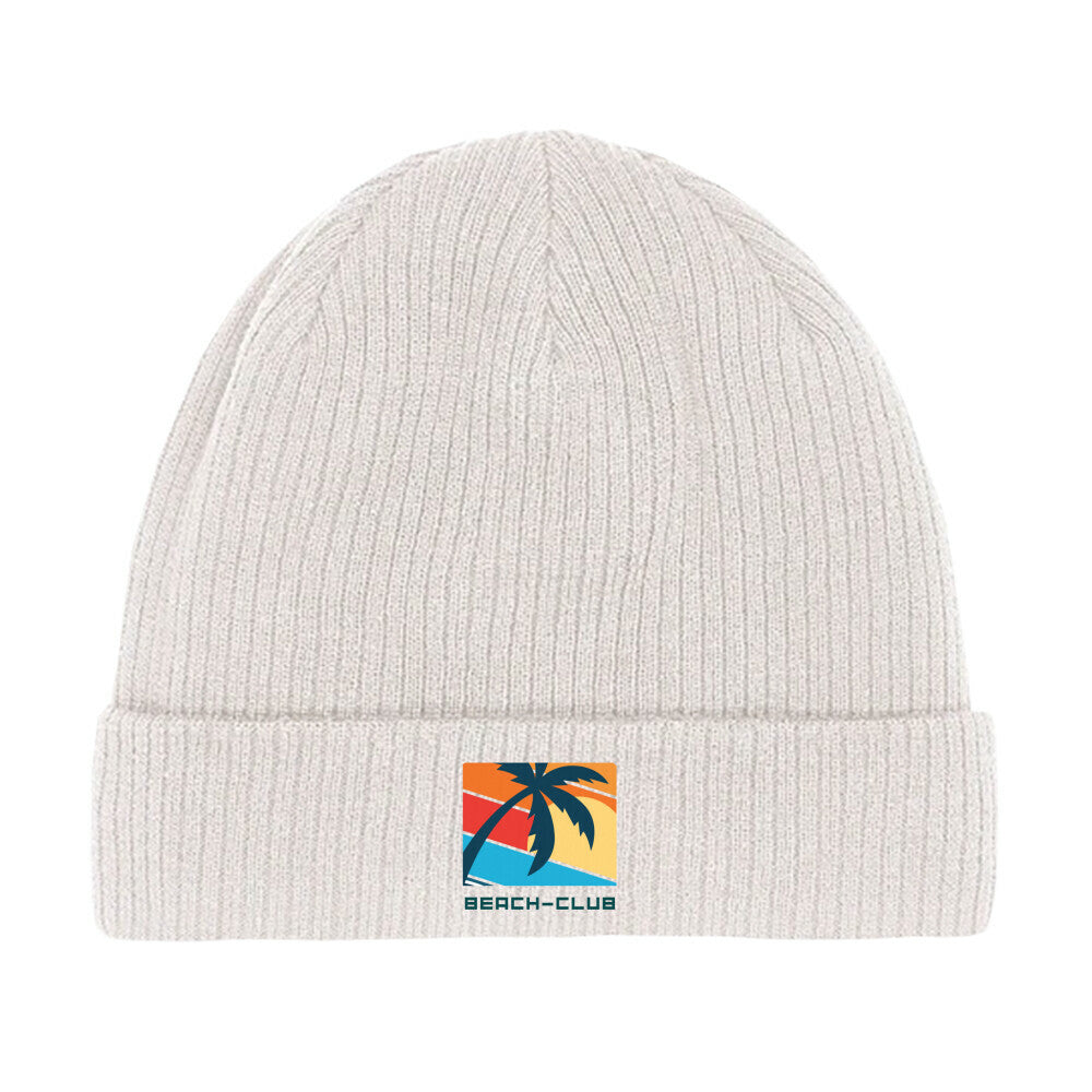 Die Beanie Mütze "Beach-Club" von MarketPrint ist aus weißer Bio-Baumwolle, doppellagig gestrickt und mit einem Patch verziert, der eine Palme, einen Sonnenuntergang und "BEACH-CLUB" in blauen Buchstaben zeigt.