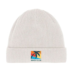 Die Beanie Mütze "Beach-Club" von MarketPrint ist aus weißer Bio-Baumwolle, doppellagig gestrickt und mit einem Patch verziert, der eine Palme, einen Sonnenuntergang und "BEACH-CLUB" in blauen Buchstaben zeigt.