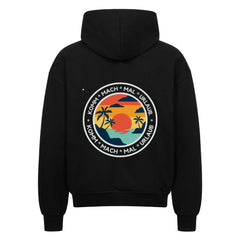 Der MarketPrint Oversized Zipper Hoodie "Urlaub" ist ein schwarzer Kapuzenpulli mit einer kreisförmigen Rückengrafik von Sonnenuntergang, Palmen und Wasser sowie "KOMM * MACH * MAL * URLAUB" am Rand. Er ist aus 100% Baumwolle gefertigt und bietet einen hohen Tragekomfort.