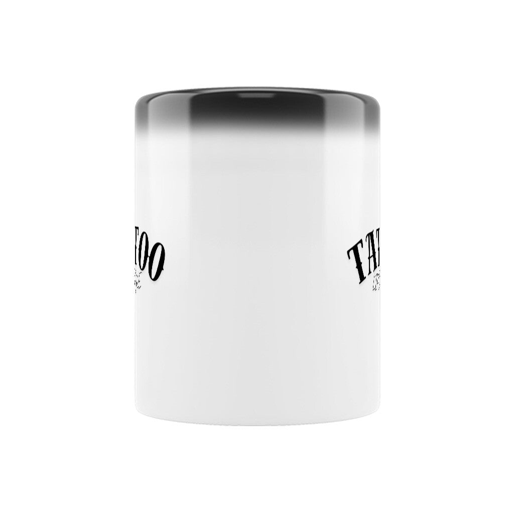 Die MarketPrint Zaubertasse "TATTOO" ist eine weiße keramische magische Tasse mit schwarzem Rand und stilisiertem schwarzem Text auf jeder Seite, die vor einem schlichten weißen Hintergrund steht.