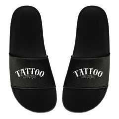 Ein Paar schwarze MarketPrint Badelatschen "Tattoo" aus PVC mit dem Schriftzug "TATTOO LIFESTYLE" in weiß auf den Trägern, die von oben auf einem schlichten weißen Hintergrund zu sehen sind.