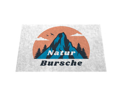 Die MarketPrint Fußmatte "Naturbursche" zeigt ein Bergmotiv mit Bäumen, Wolken und Vögeln vor einer orangen Sonne sowie dem Schriftzug "Natur Bursche" - strapazierfähig und schmutzabweisend.