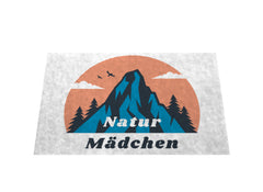 Die MarketPrint Fußmatte "Natur Mädchen" zeigt einen stilisierten blauen Berg, Tannenbäume, eine orangefarbene Sonne und fliegende Vögel mit dem Schriftzug Natur Mädchen darunter - ideal für den Eingang, um Schmutz fernzuhalten.